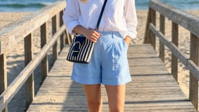Relaxed family picture outfits summer. A woman in a crisp white oversized button-down shirt, sky blue tailored linen shorts, off-white espadrille flats, and a nautical stripe bag for a beach photoshoot.