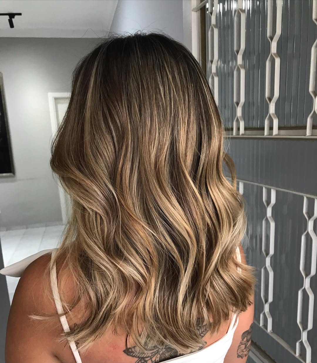 Woman with golden balayage summer 2026 hair wearing a flowy white sundress, sunglasses, and sandals, enjoying a sunny day outdoors.