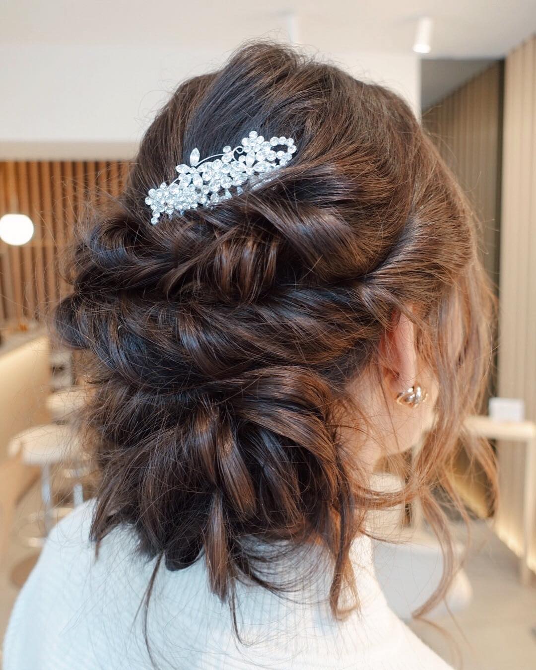 Beautiful blonde woman with a soft, romantic updo, perfect outdoor wedding hair for summer 2026. Her hair is loosely pinned with delicate curls framing her face, adorned with small white floral accents, ideal for a garden or rustic wedding.