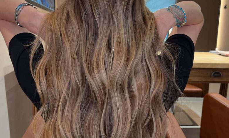 Woman with long wavy hair featuring sun-kissed summer babylights balayage, glowing softly against a blurred outdoor backdrop.