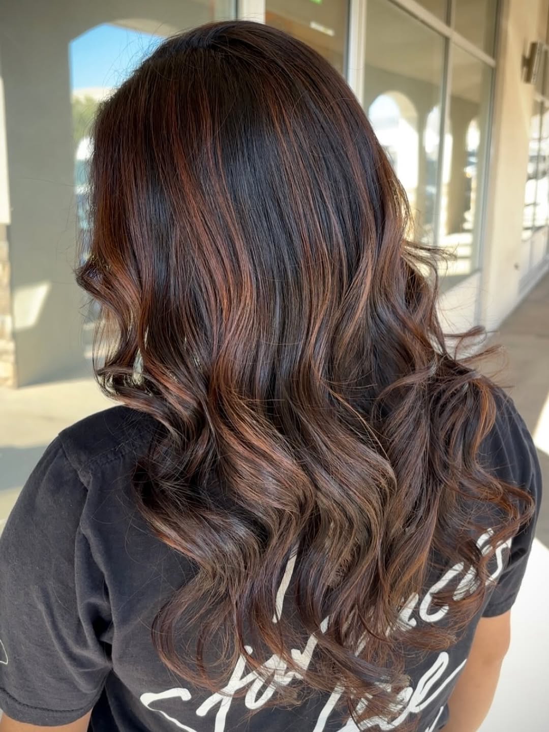 Stunning Caramel Brown Summer Hair Looks 2026 Woman with long wavy caramel brown summer hair, wearing a white off-the-shoulder top and jeans, casually posing outdoors for a summer look.
