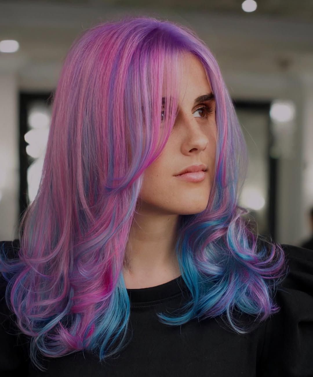 Close-up of a woman with vibrant pastel purple and pink hair, styled in soft waves, against a blurred outdoor summer background. Ideal for pastel hair color summer 2026 inspiration.