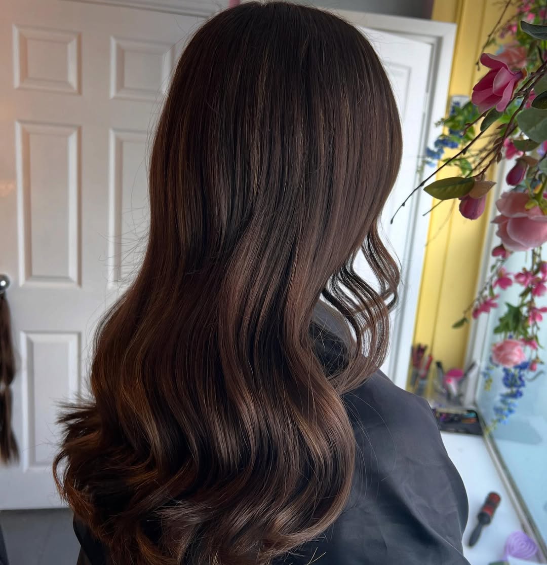 Stunning Wavy Summer Hair for Brunettes 2026 A brunette woman with long, shiny, wavy summer hair for brunettes 2026, styled casually over one shoulder, wearing a white off-the-shoulder top.